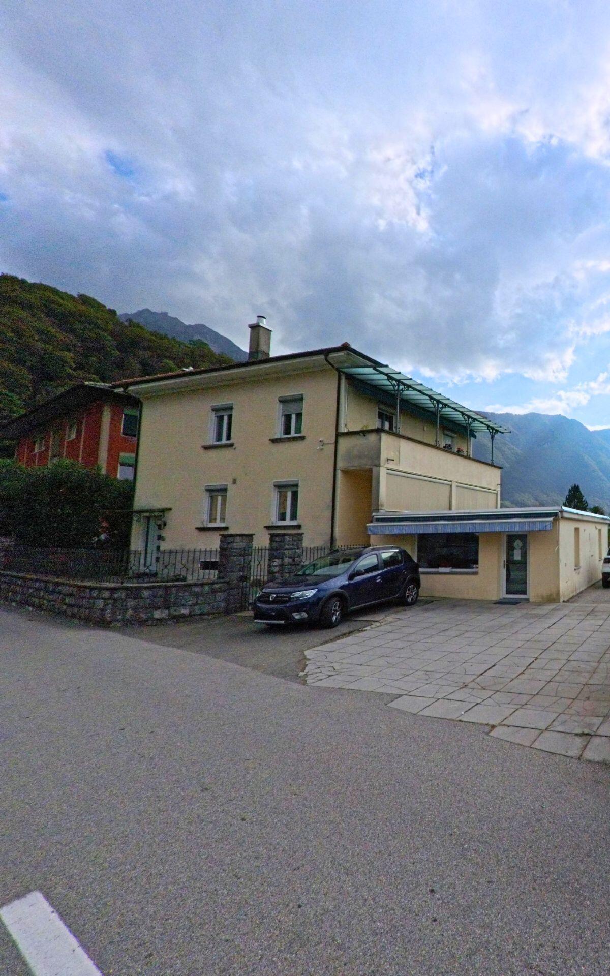 Ein beiges Wohnhaus mit ueberdachter Terrasse und kleinem Anbau steht in Val Mara (6818, Kanton Tessin) vor einer beeindruckenden Bergkulisse.