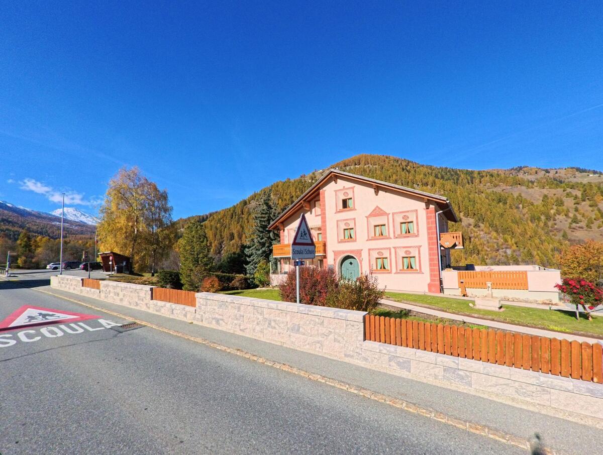 An einer ruhigen Dorfstrasse in Val Müstair (7536, Kanton Graubünden) steht ein gepflegtes Schulhaus mit dekorierter Fassade vor herbstlich gefärbten Berghängen.