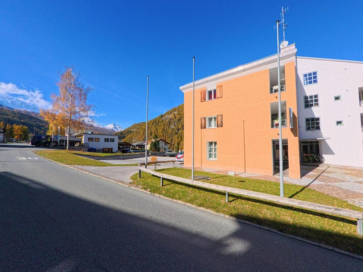 Neben einer ruhigen Quartierstrasse steht ein modernes Wohngebäude in Val Müstair (7536, Kanton Graubünden), während sich dahinter bewaldete Berghänge und schneebedeckte Gipfel abzeichnen.