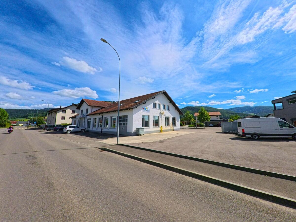 Im Industriegebiet von Val Terbi (2824, Kanton Jura) steht ein weisses Gewerbegebäude mit roten Ziegeldächern unter einem weit geöffneten Sommerhimmel.