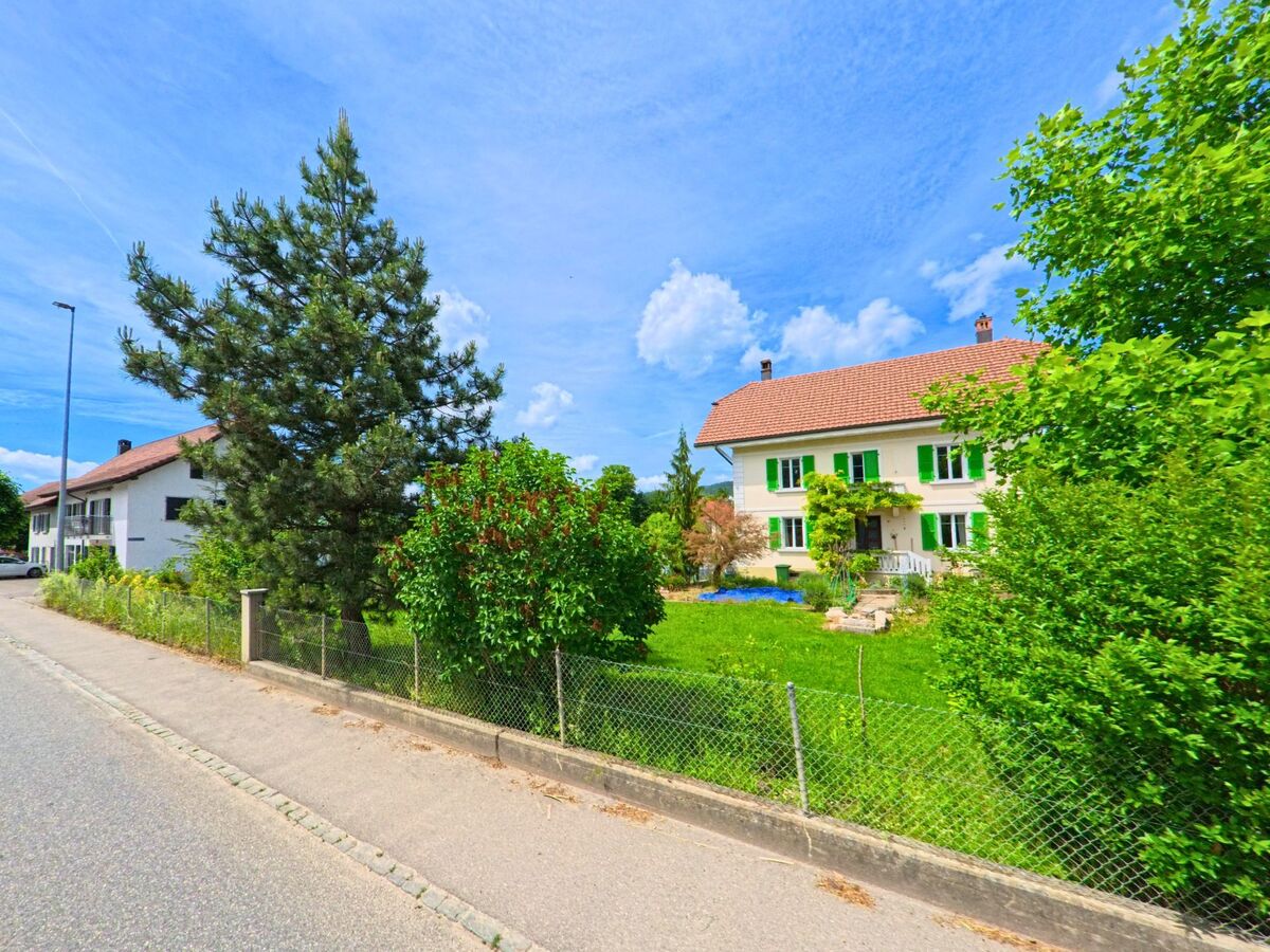 Ein gepflegtes Haus mit grünen Fensterläden steht in Val Terbi (2824, Kanton Jura) hinter einem blühenden Garten und hohen Bäumen.