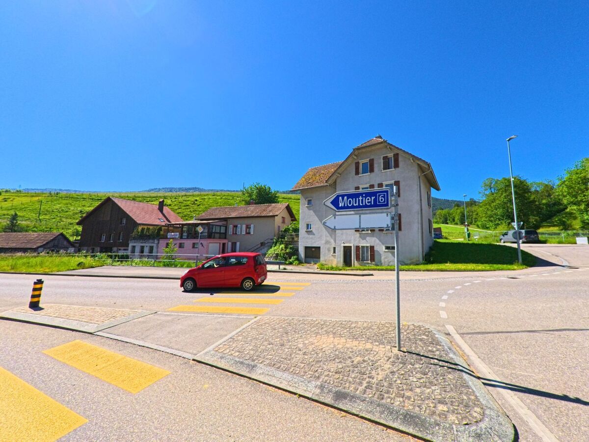 In Valbirse (PLZ 2733, Kanton Bern) zeigt ein Wegweiser an einer Kreuzung die Richtung nach Moutier, während ein rotes Auto den Fussgängerstreifen überquert.