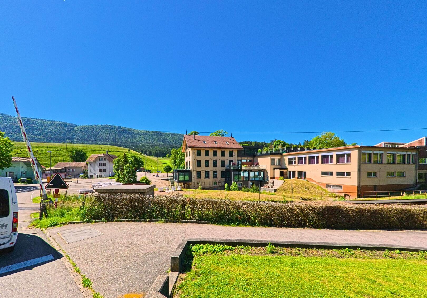 Im Zentrum von Valbirse (PLZ 2733, Kanton Bern) stehen moderne Schulgebäude und ein renoviertes Altbauhaus vor bewaldeten Jurahängen.