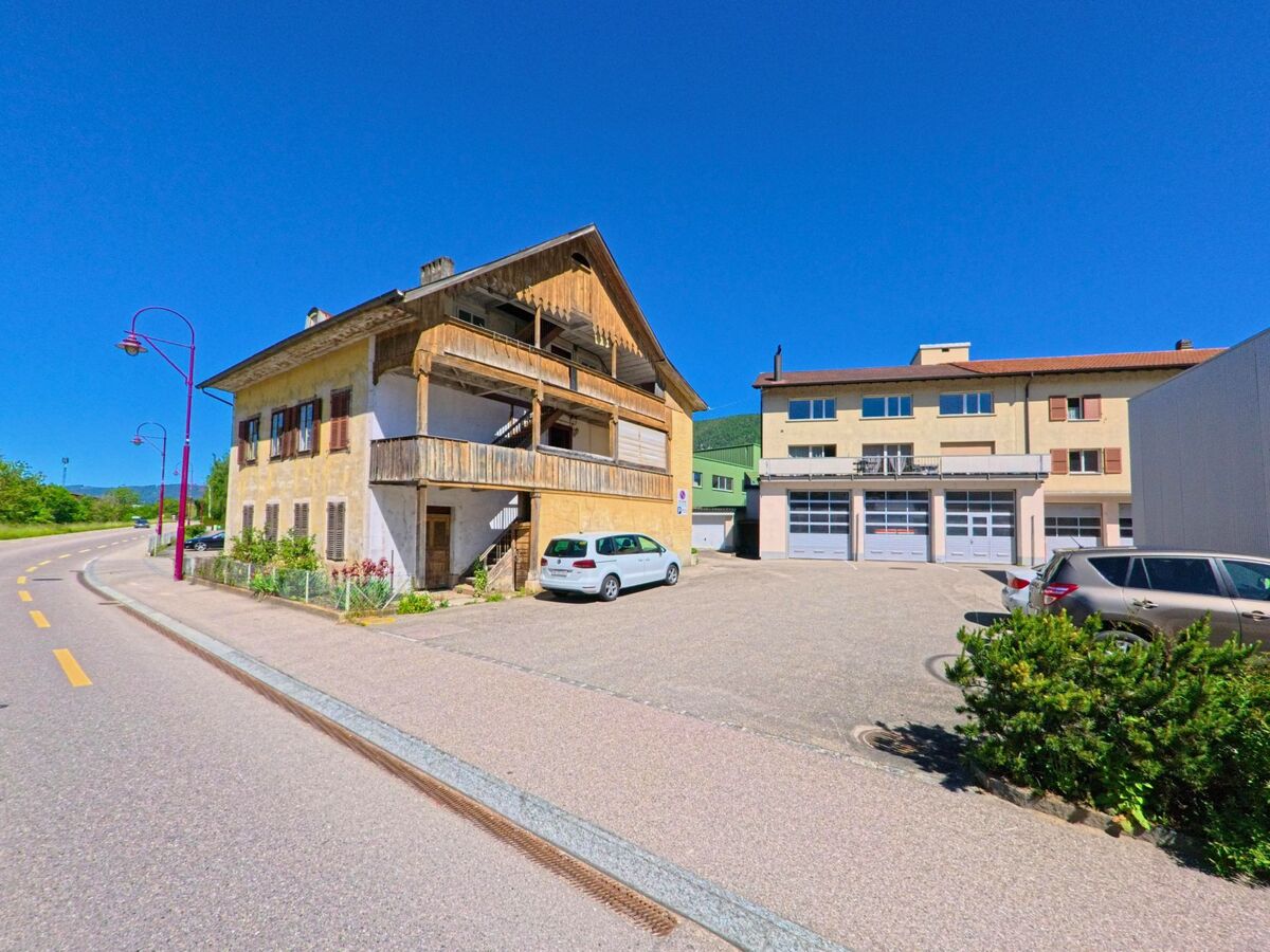 In Valbirse (2735, Kanton Bern) stehen ein renovierungsbedürftiges Holzhaus und ein modernes Gebäude mit Garagentoren nebeneinander an einer leicht geschwungenen Strasse.