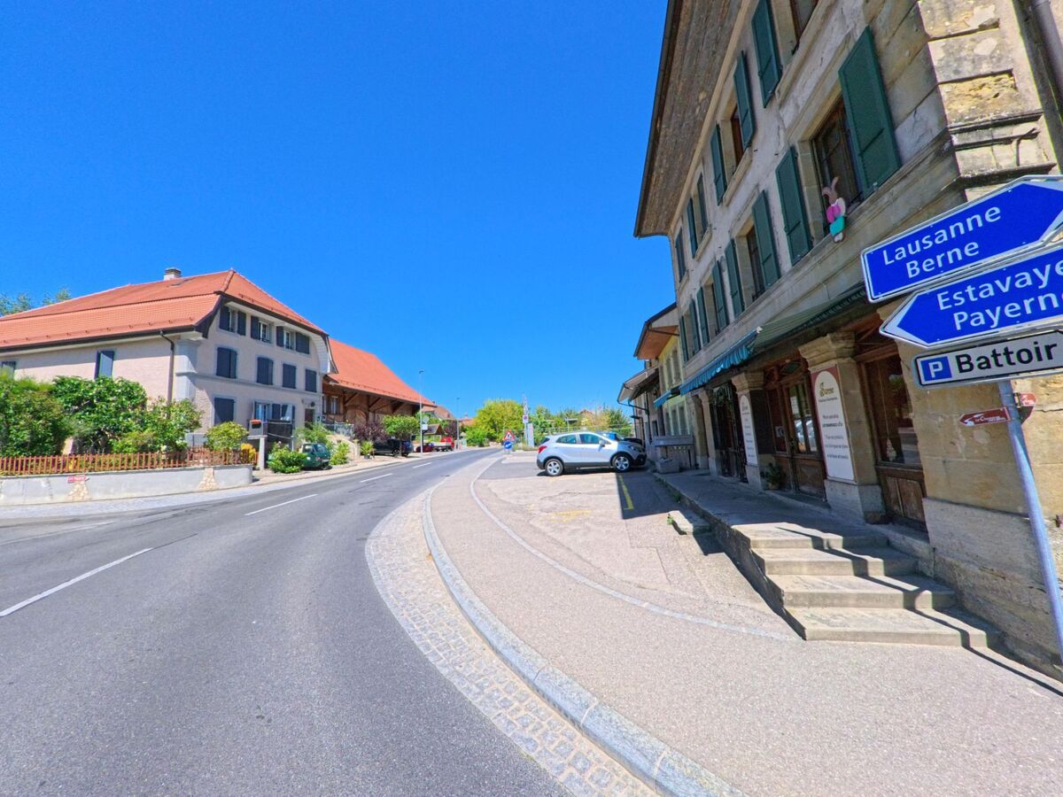 Eine leicht ansteigende Strasse in Valbroye (1523, Kanton Waadt) führt an traditionellen Häusern mit roten Dächern und Wegweisern vorbei.