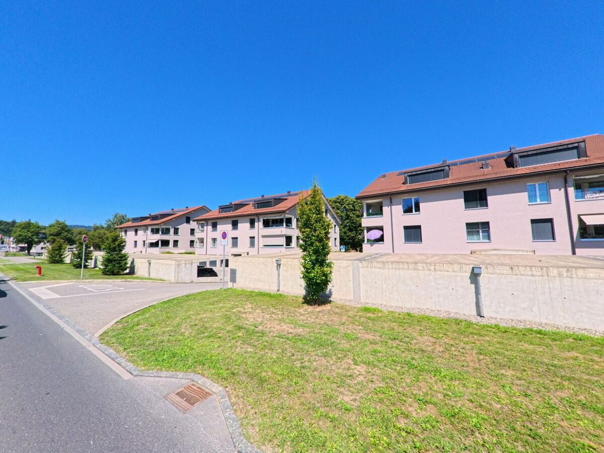 Mehrfamilienhaeuser mit roten Daechern stehen in Valbroye (1523, Kanton Waadt) entlang einer ruhigen Strasse mit gruener Wiese im Vordergrund.