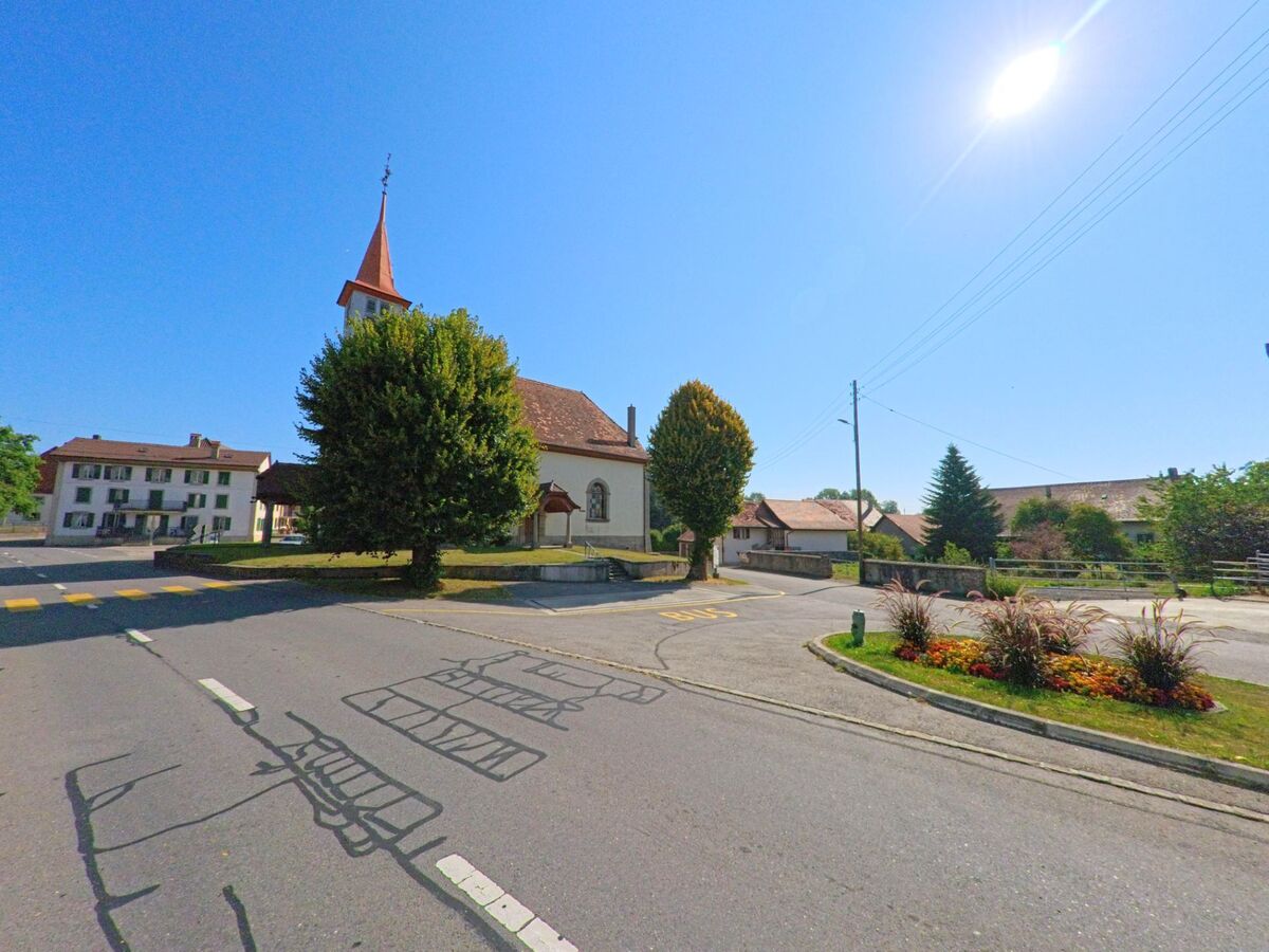 Eine Dorfkirche mit rotem Turm steht in Valbroye (1535, Kanton Waadt) an einer ruhigen Kreuzung mit Bäumen und gepflegtem Strassenrand.