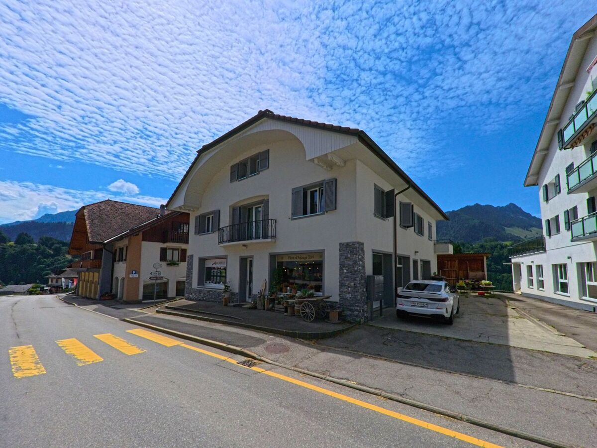 Im Ortskern von Val-de-Charmey (1647, Kanton Freiburg) steht ein modernes Wohn- und Geschäftshaus mit Ladenlokal an einer Strasse mit Zebrastreifen, flankiert von weiteren Gebäuden vor Bergkulisse.