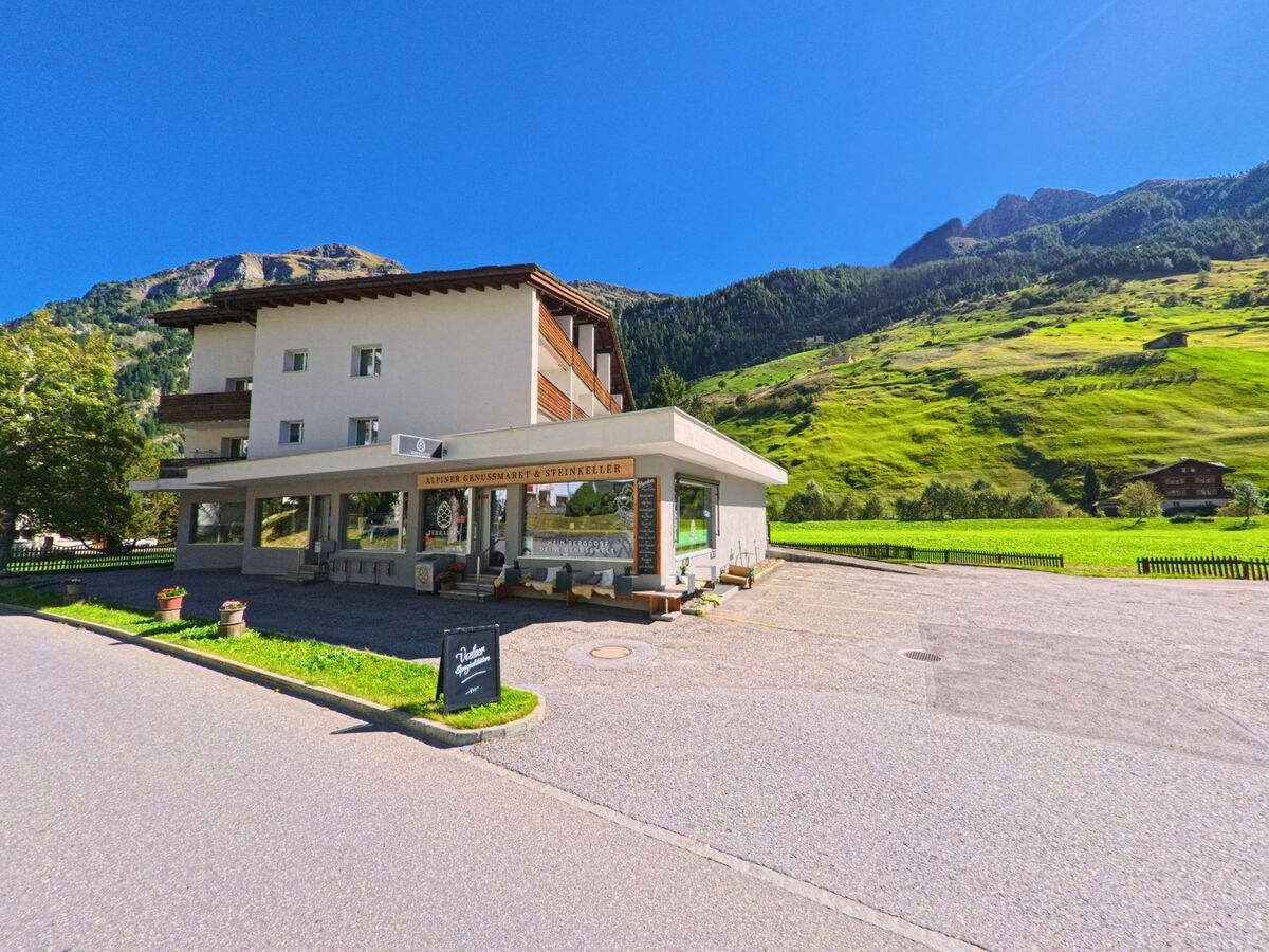 In Vals (7132, Kanton Graubünden) steht ein modernes Gebäude mit dem Alpen Genussmarkt vor einer weiten, grünen Bergwiese unter klarem Himmel.