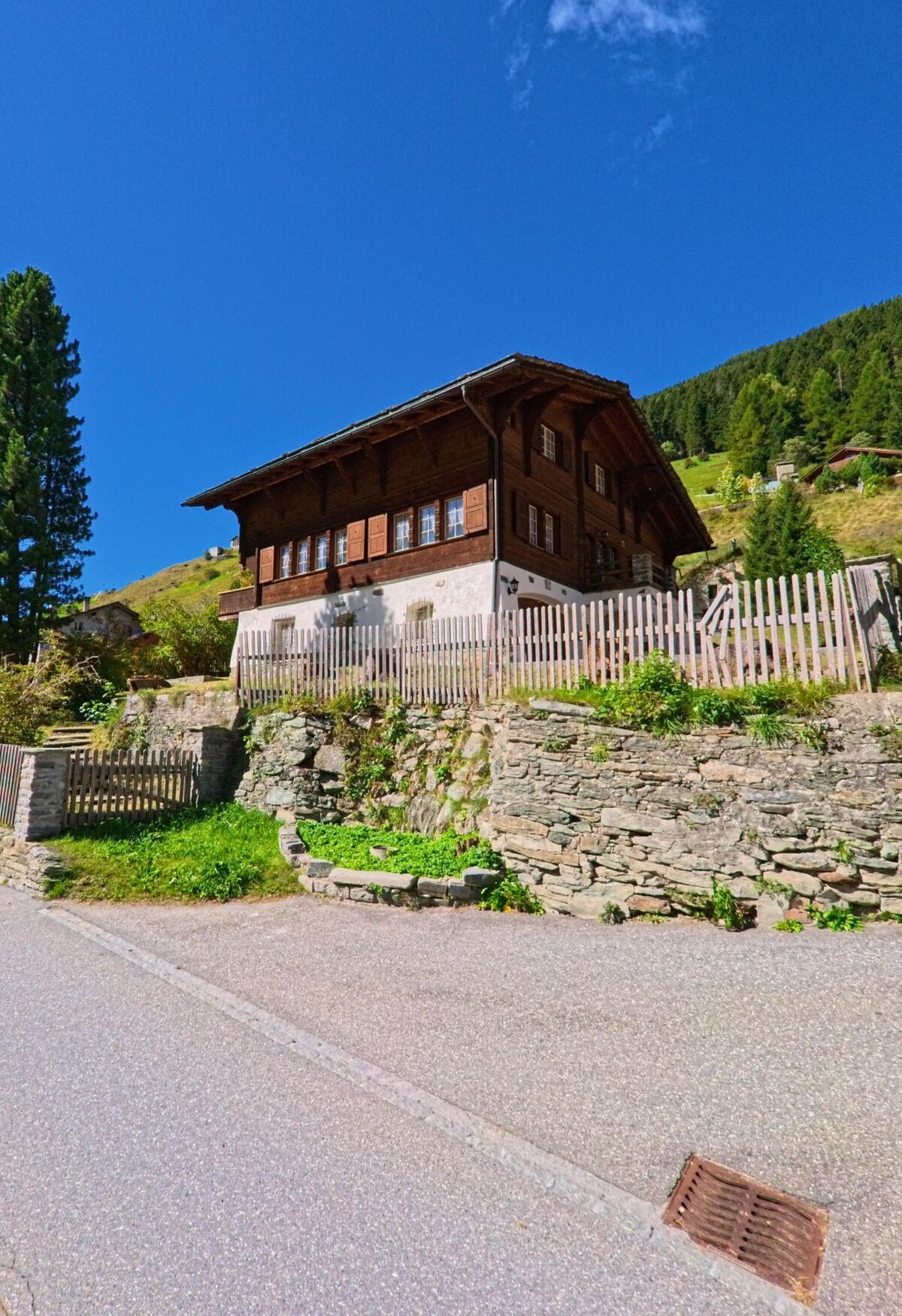 Ein traditionelles Chalet aus Holz steht in Vals (7132, Kanton Graubünden) auf einer kleinen Anhöhe mit Natursteinmauer und Blick auf bewaldete Berghänge.