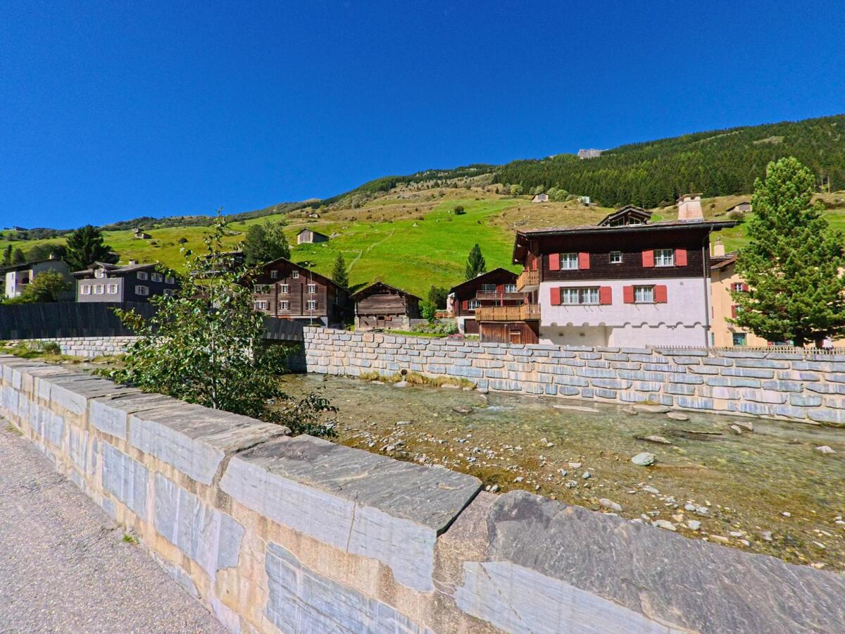 In Vals (7132, Kanton Graubünden) verlaufen traditionelle Holzhäuser mit roten Fensterläden entlang eines klaren Bachs vor den grünen Hängen des Tals.