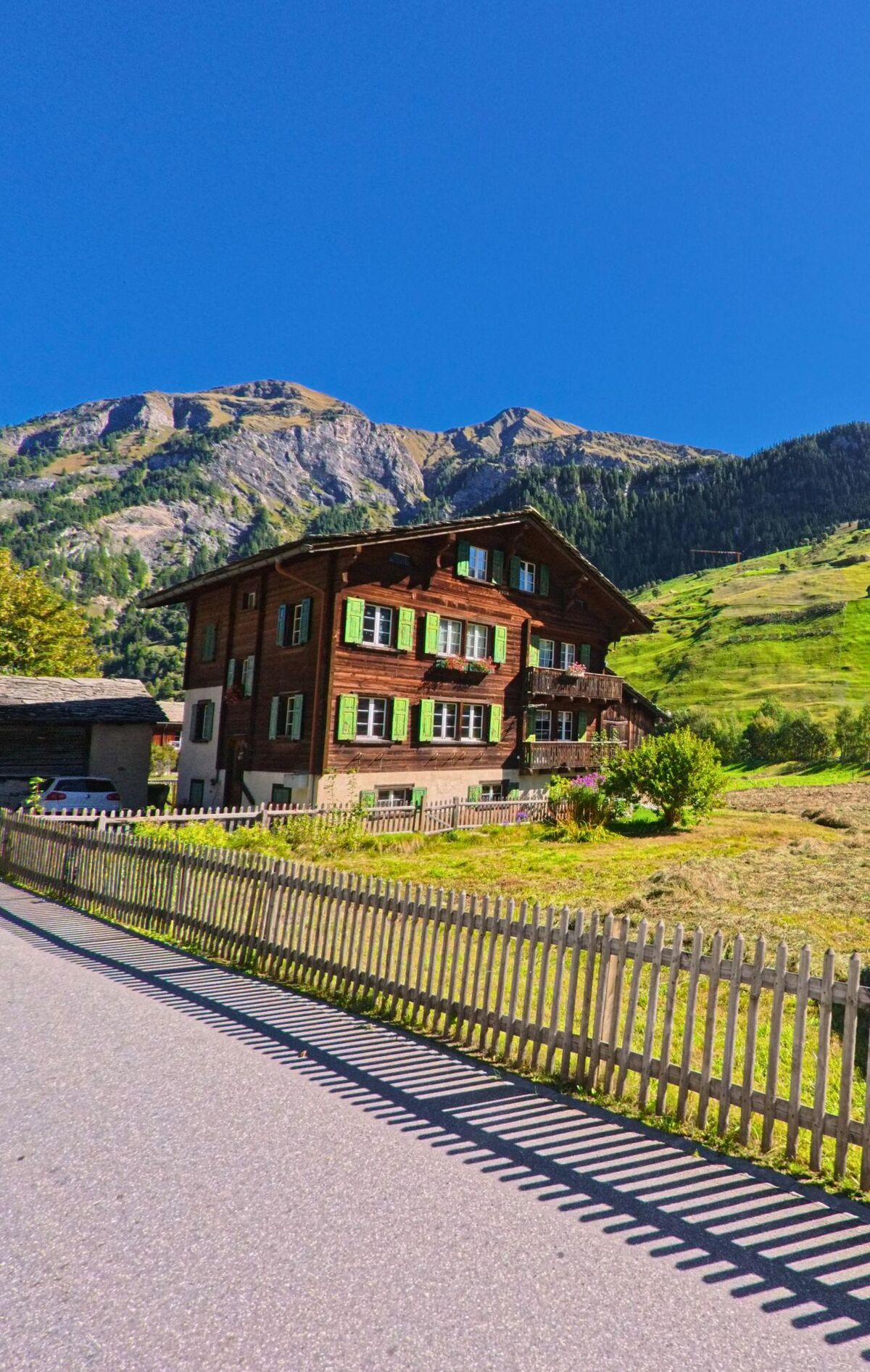 Ein traditionelles Holzhaus mit grünen Fensterläden steht in Vals (7132, Kanton Graubünden) am Rand einer Wiese vor den steilen Hängen der umliegenden Berge.