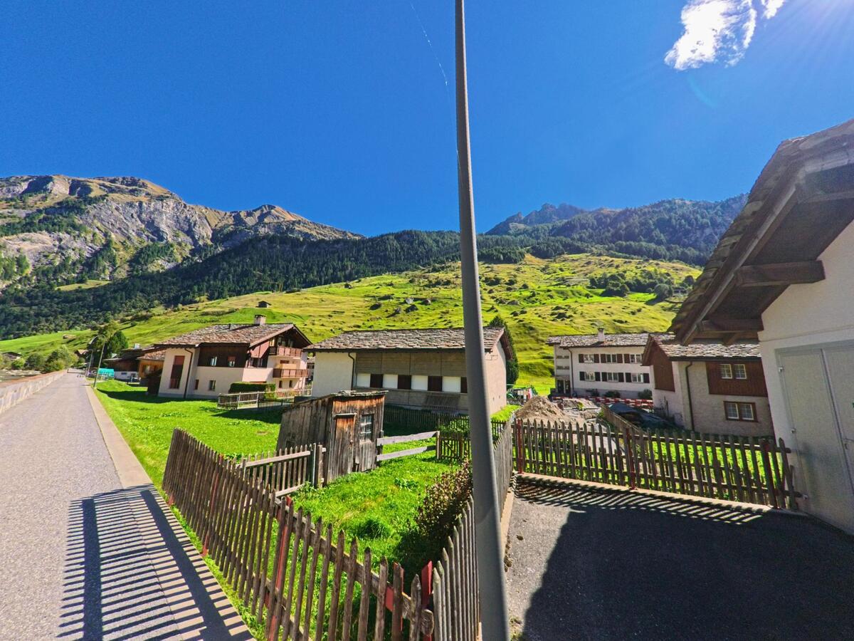 Entlang einer ruhigen Strasse in Vals (7132, Kanton Graubünden) stehen Wohnhäuser mit Holzzäunen vor den steilen, grünen Hängen der Berge.