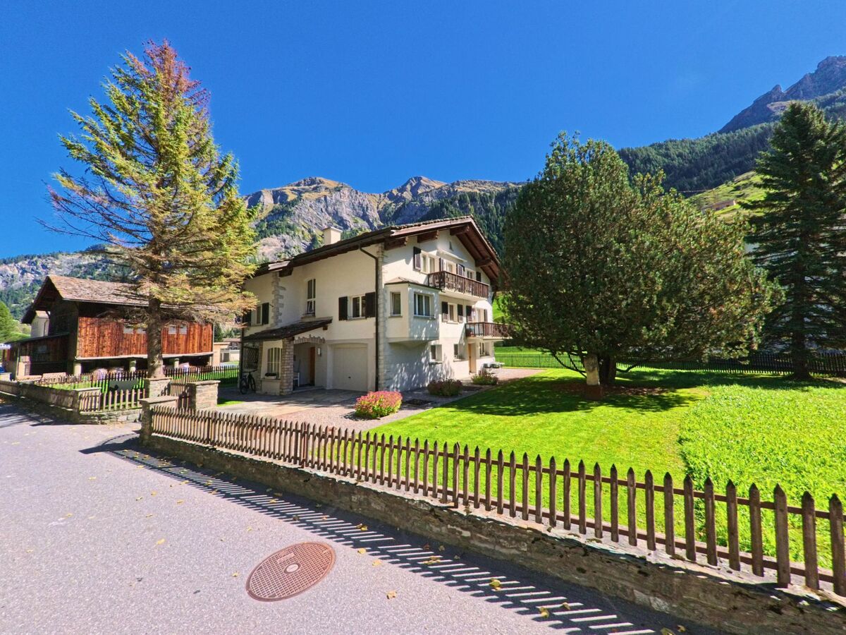 Ein Wohnhaus mit gepflegtem Garten und Holzzaun steht in Vals (7132, Kanton Graubünden) vor einer eindrucksvollen Bergkulisse unter klarem Himmel.