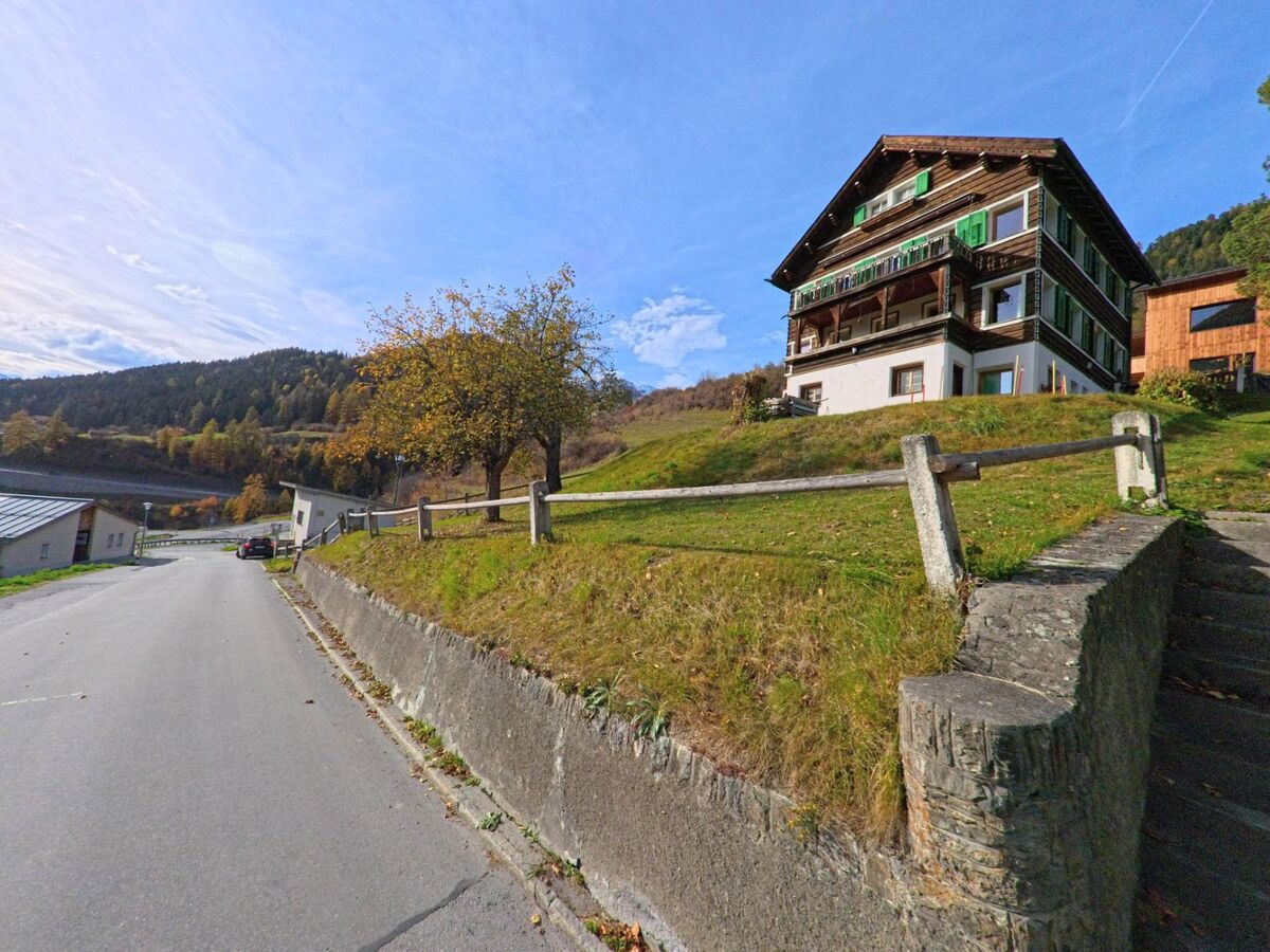 Am Hang oberhalb einer ruhigen Strasse steht ein traditionelles Wohnhaus in Valsot (7556, Kanton Graubünden), umgeben von Wiesen und herbstlich gefärbten Bäumen.