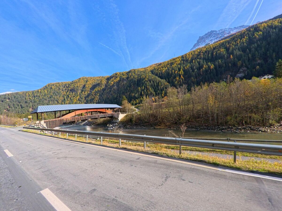 Entlang der Talstrasse bei Valsot (7559, Kanton Graubünden) überspannt eine gedeckte Holzbrücke den Fluss, während sich dahinter herbstlich gefärbte Waldhänge erheben.