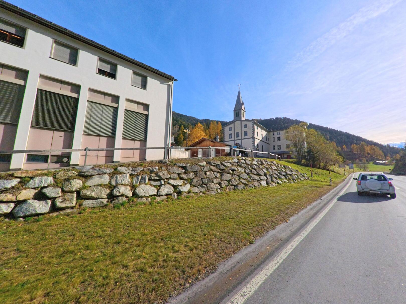 Entlang der Hauptstrasse von Valsot (7560, Kanton Graubünden) liegen öffentliche Gebäude und eine Kirche auf einer erhöhten Geländestufe vor herbstlich gefärbten Hängen.