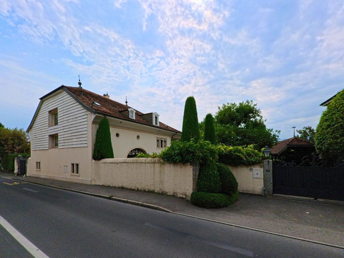 Ein grosses Wohnhaus mit verziertem Dach und gepflegtem Garten liegt in Vandoeuvres (1253, Kanton Genf) direkt an der Strasse hinter einer hohen Mauer.