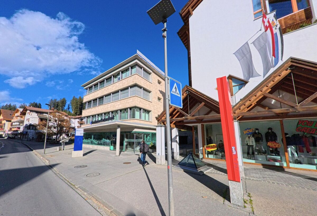 Entlang einer belebten Strasse mit Geschäften und einem Bankgebäude zeigt sich ein zentraler Abschnitt von Vaz/Obervaz (7078, Kanton Graubünden) bei klarem Himmel.