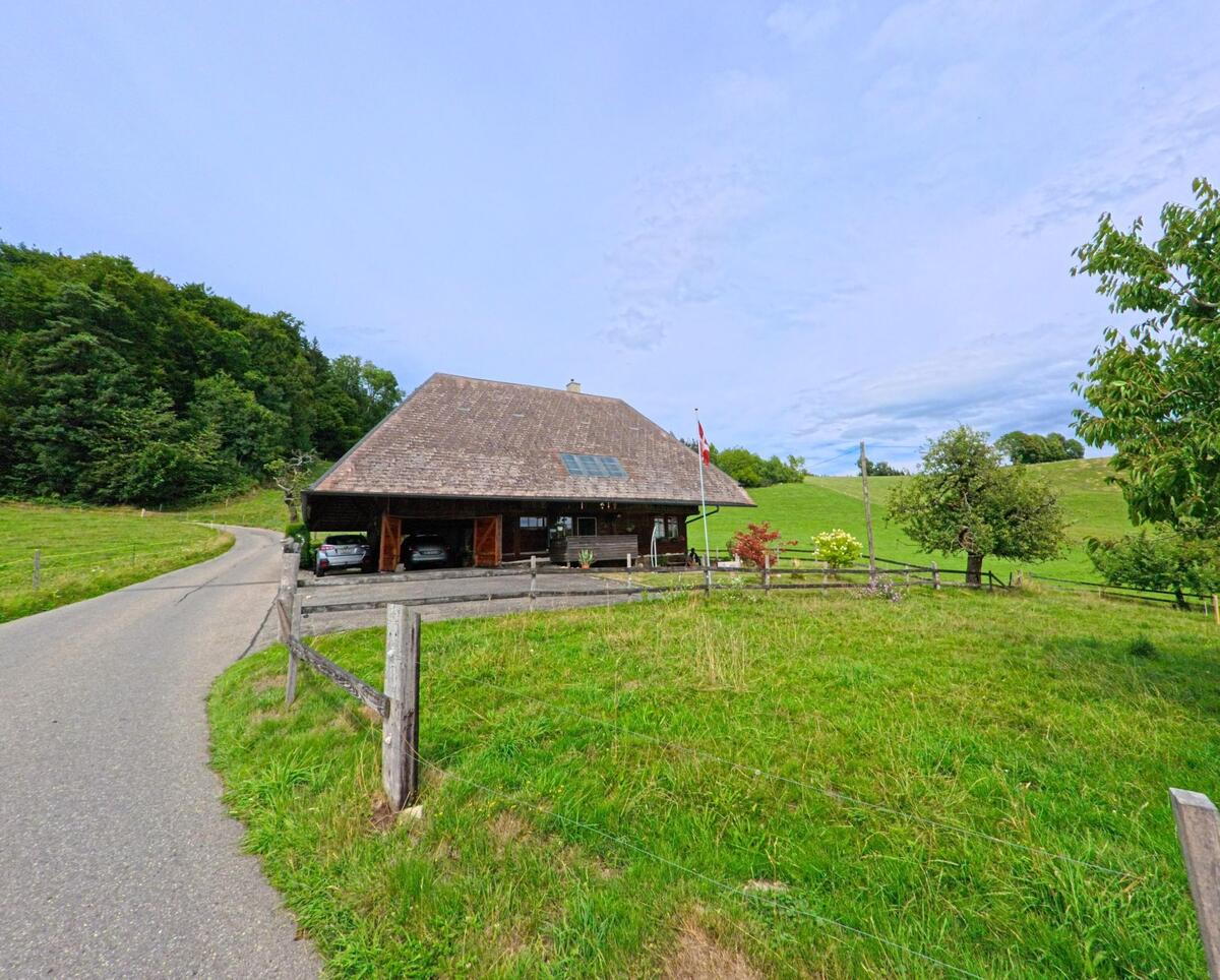 Ein abgelegenes Bauernhaus mit grossem Walmdach steht in Vechigen (3067, Kanton Bern) an einer schmalen Strasse, umgeben von Weiden und sanften Huegeln.