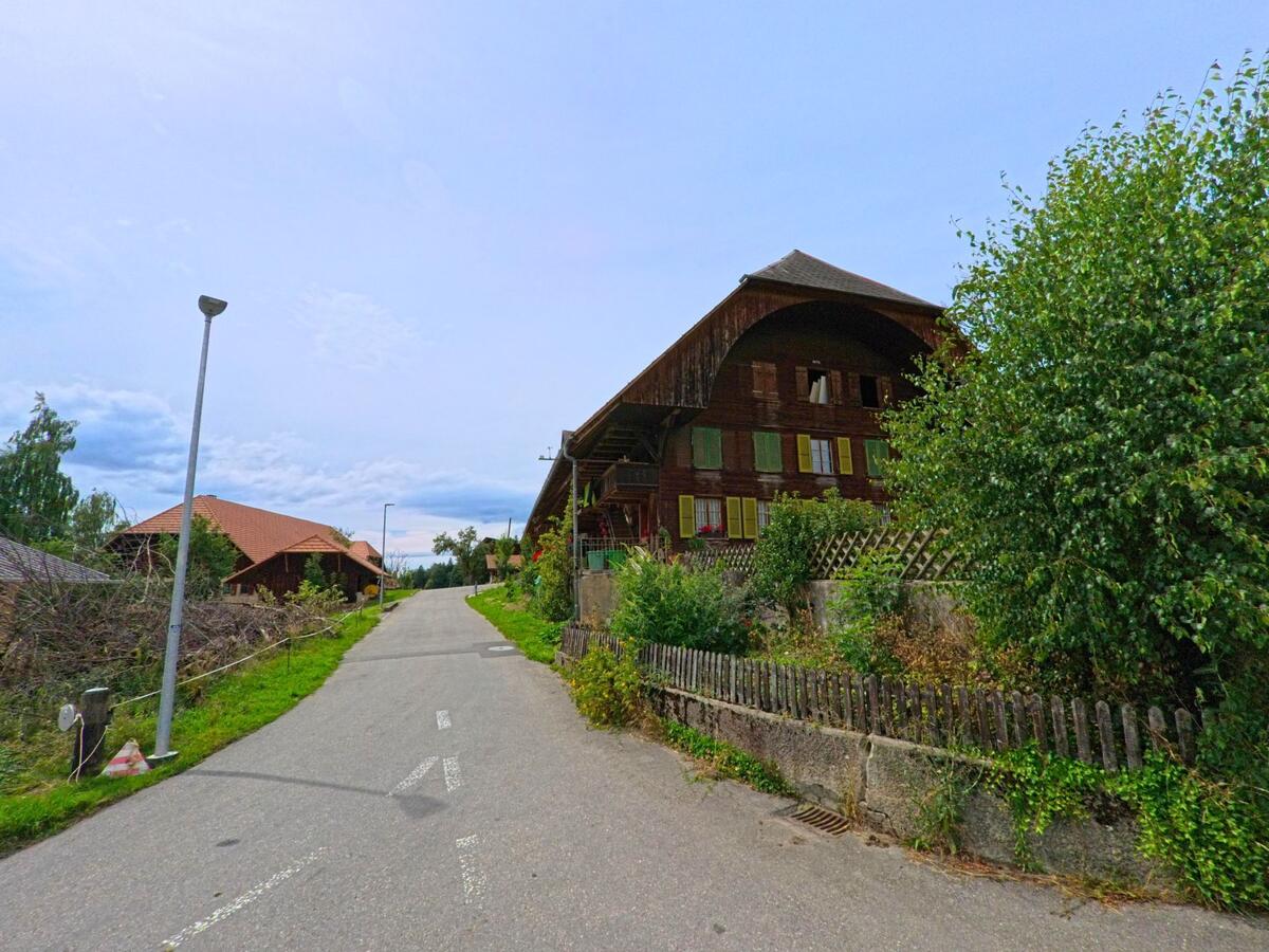 Entlang einer schmalen Strasse in Vechigen (3067, Kanton Bern) steht ein traditionelles Bauernhaus mit gruenen Fensterlaeden und grossem Holzdach.
