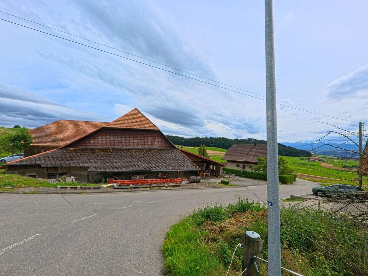 An einer Kreuzung in Vechigen (3067, Kanton Bern) steht ein grosser Bauernhof mit weit ausladendem Dach vor einer offenen Huegellandschaft.