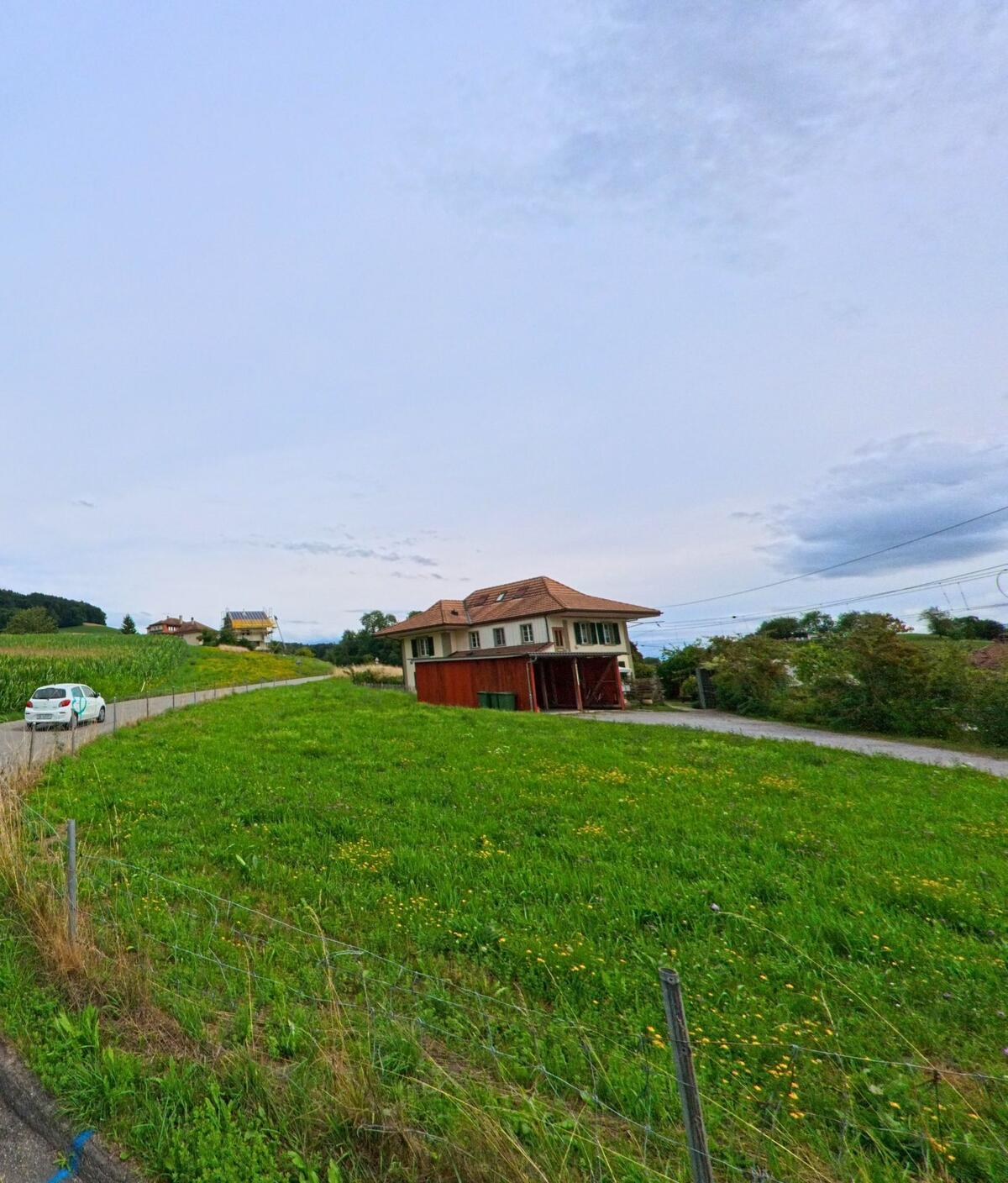 Am Rand einer schmalen Strasse in Vechigen (3067, Kanton Bern) steht ein zweistoeckiges Wohnhaus mit Nebengebaeude vor einer weiten Gruenflaeche.