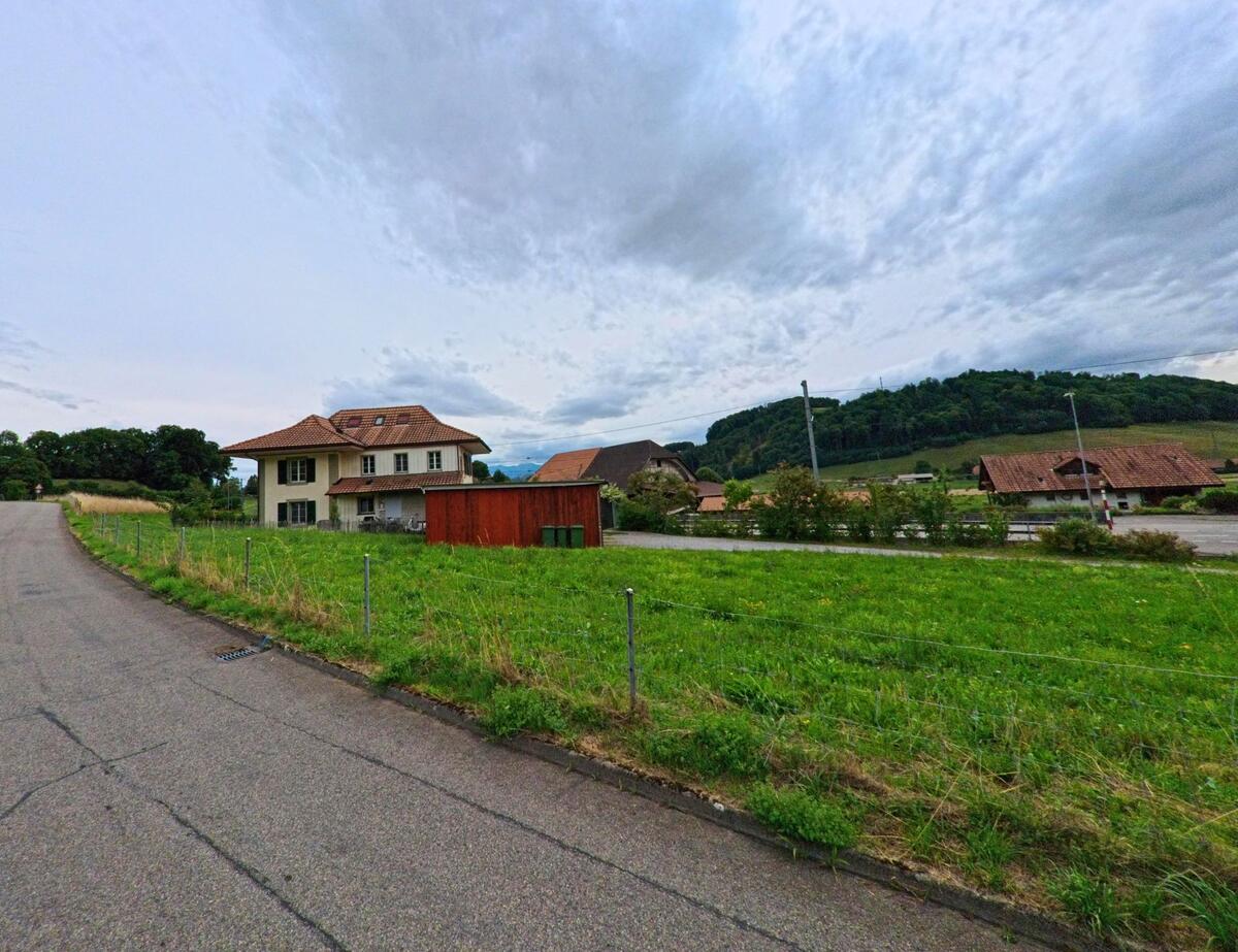 Ein helles Wohnhaus mit rotem Ziegeldach steht in Vechigen (3067, Kanton Bern) am Rand einer schmalen Strasse mit Blick auf bewaldete Huegel.