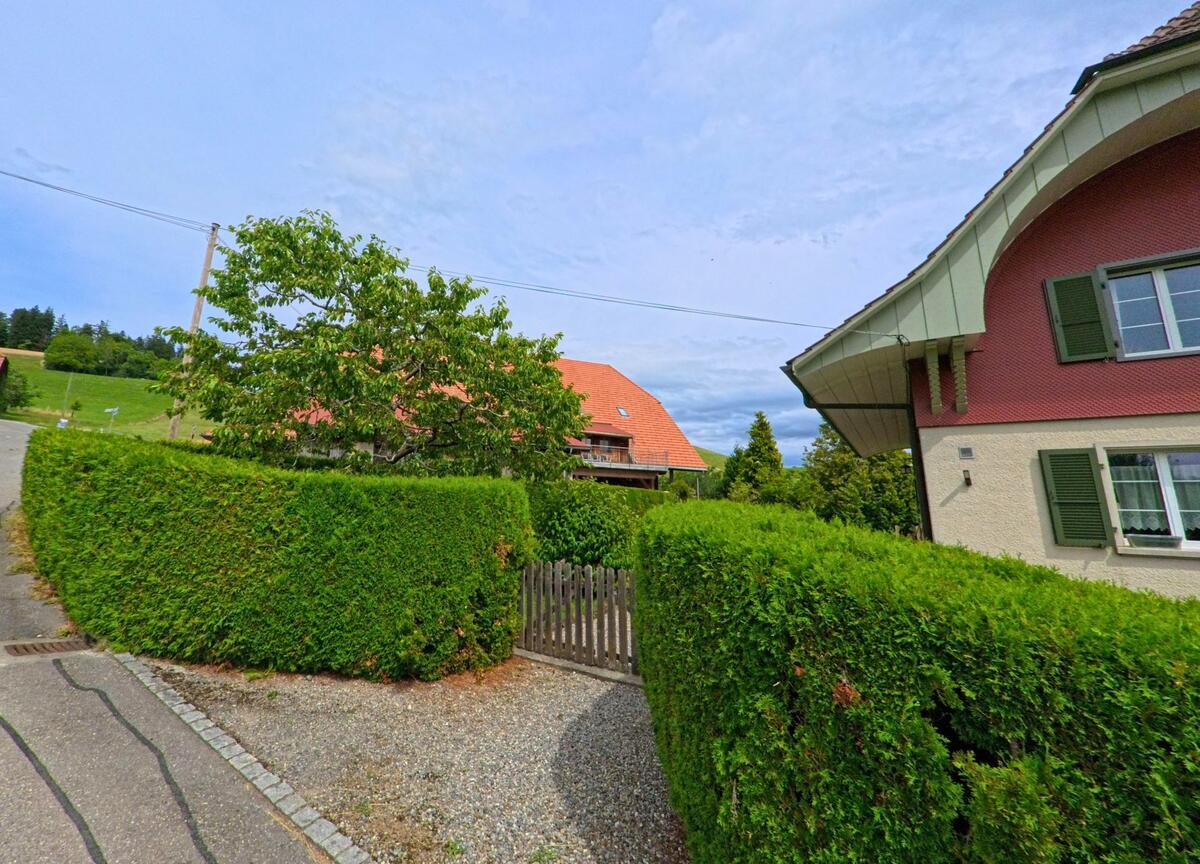 Hinter einer dichten Hecke in Vechigen (3067, Kanton Bern) ragt das rote Dach eines traditionellen Wohnhauses ueber einen Gartenzaun.