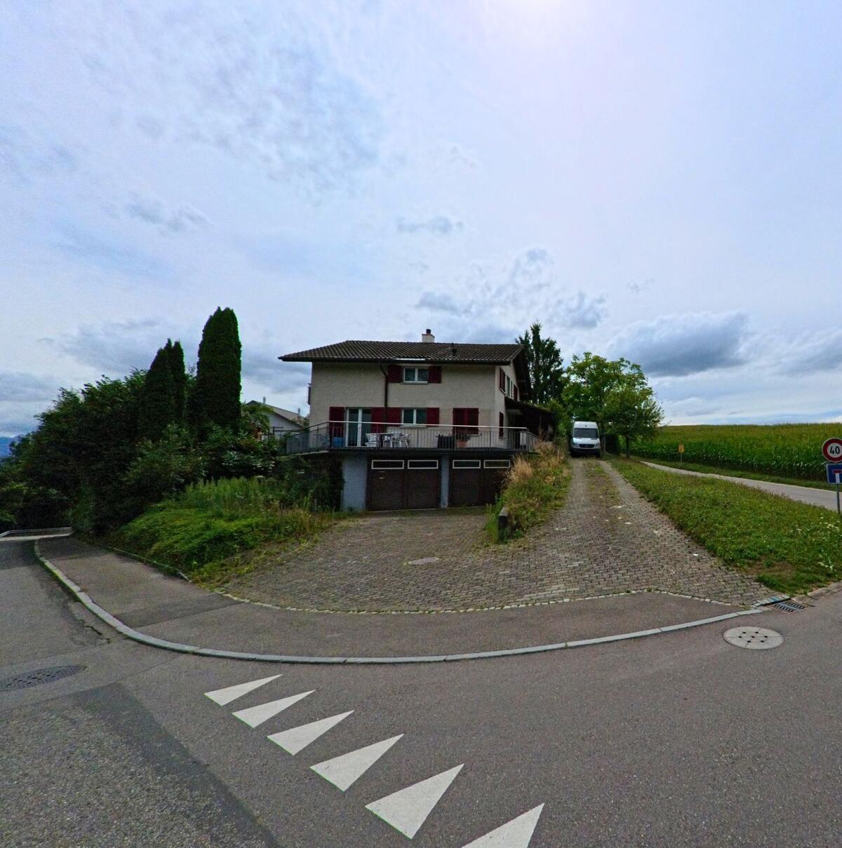 An einer Strassenkreuzung in Vechigen (3067, Kanton Bern) steht ein zweigeschossiges Wohnhaus mit roten Fensterlaeden und grosser Einfahrt.