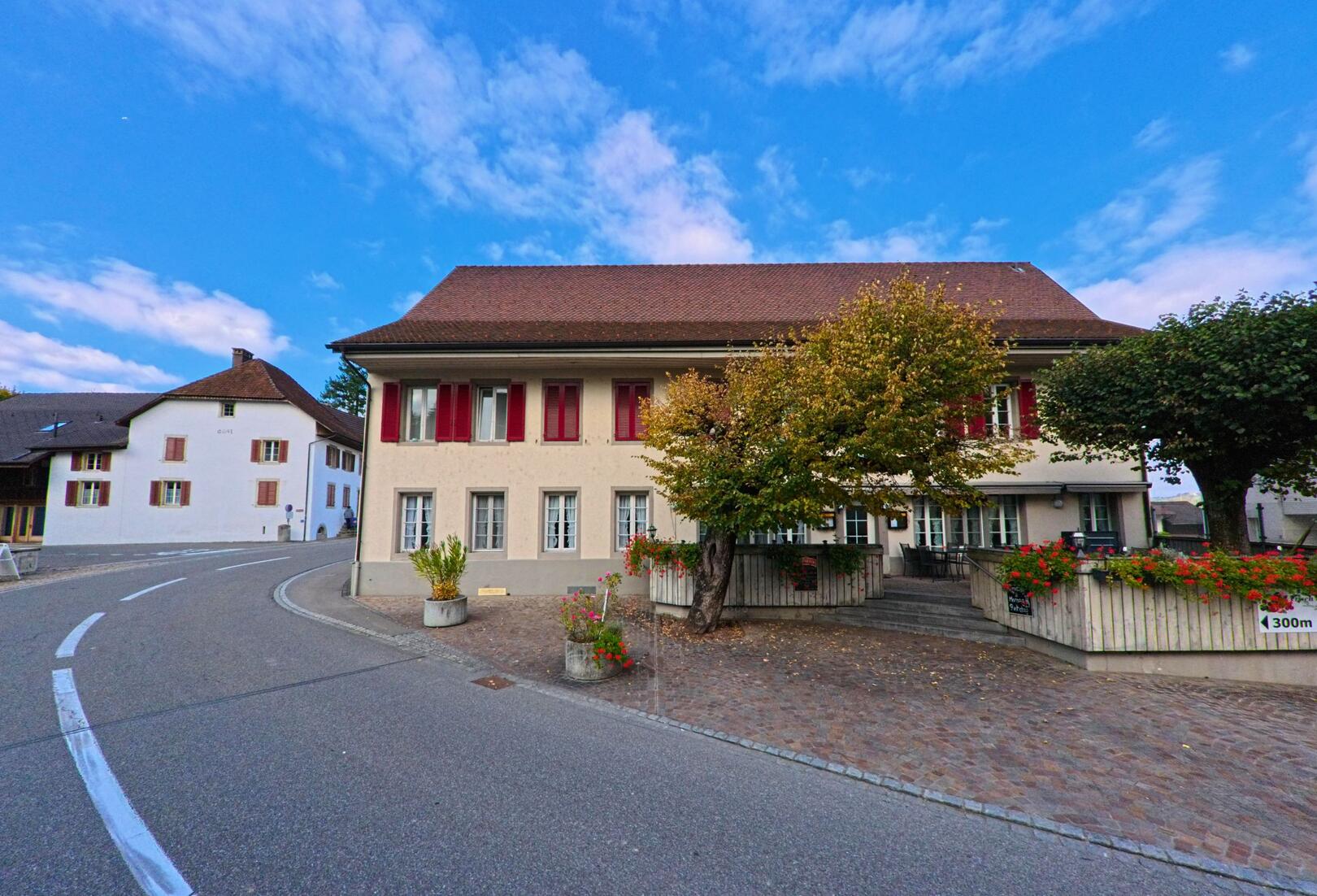 In Veltheim (5106, Kanton Aargau) praegen ein grosses Wohngebaeude mit roten Fensterlaeden und eine geschwungene Strasse das Ortsbild, begleitet von Baeumen und Blumenschmuck.