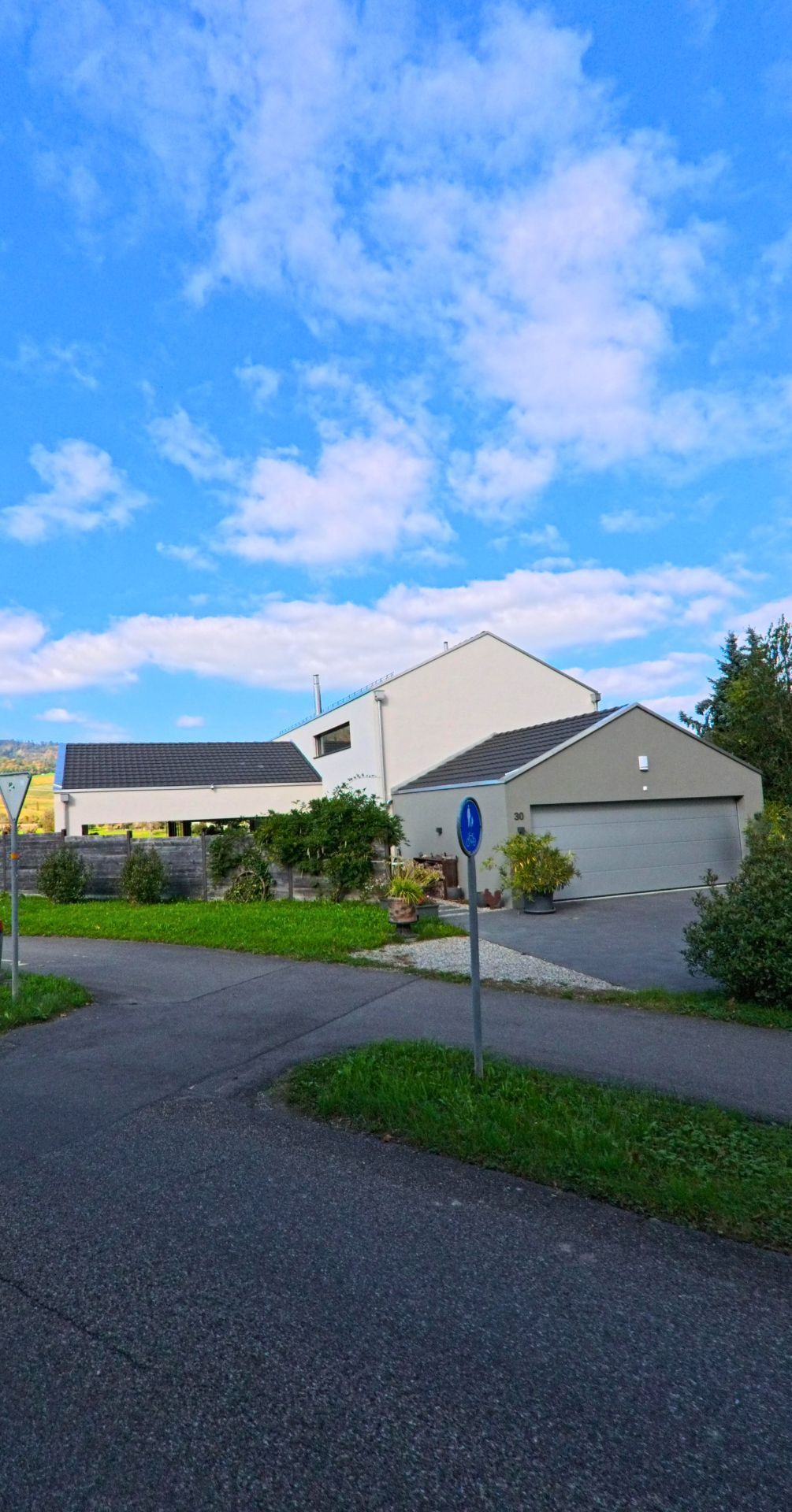 In Veltheim (5106, Kanton Aargau) steht ein modernes Wohnhaus mit grosser Garage an einer ruhigen Strassenkreuzung, umgeben von viel Gruen und vereinzelten Baeumen.