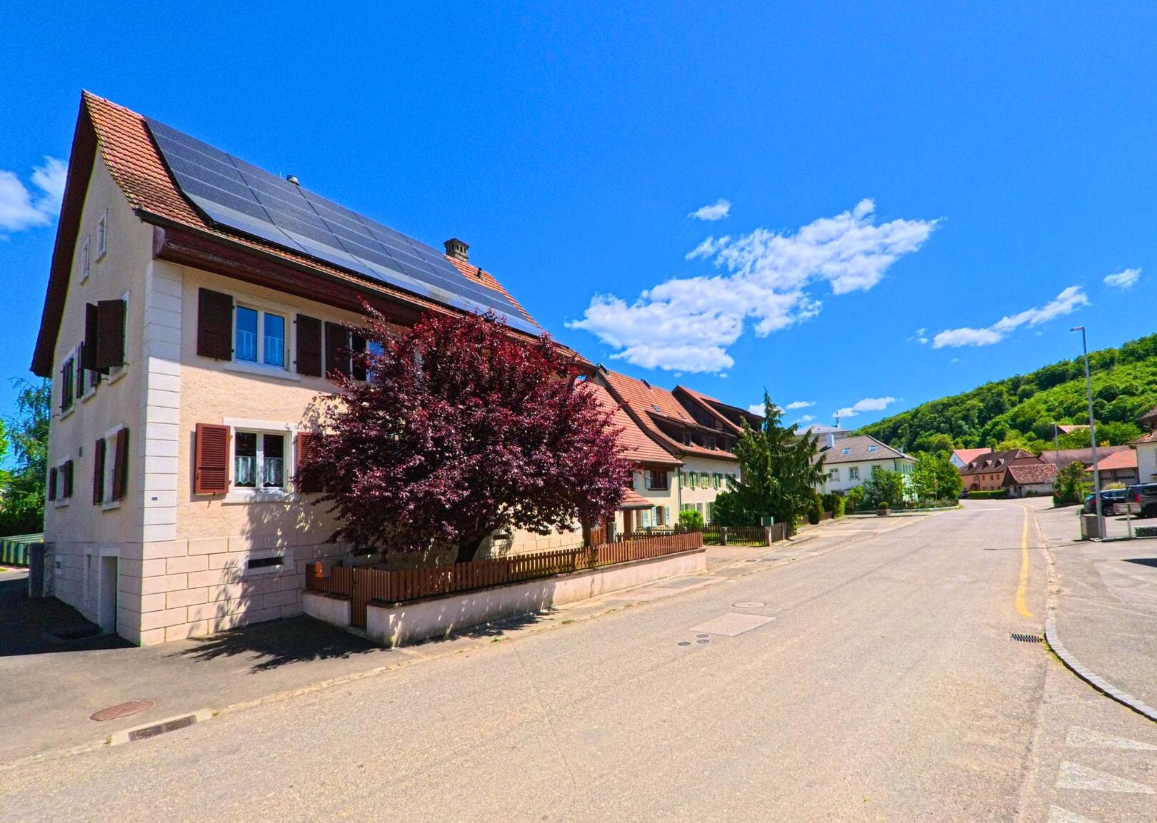 In Vendlincourt (2943, Kanton Jura) führt eine Strasse entlang eines Hauses mit einer Solaranlage auf dem Dach und einem grossen Baum im Vordergrund. Die umliegende Landschaft ist grün und hügelig.