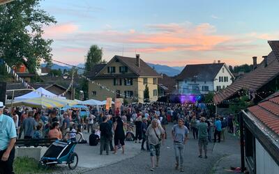 Bei einem Dorffest in Hausen am Albis (8915, Kanton Zürich) versammeln sich viele Menschen zwischen Zelten und beleuchteten Ständen in der Abenddämmerung.