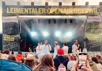 Beim Leimentaler OpenAir in Oberwil (4104, Kanton Baselland) genießen Besucher das Konzert einer Band, während eine Zuschauerin das Ereignis mit ihrem Handy filmt.