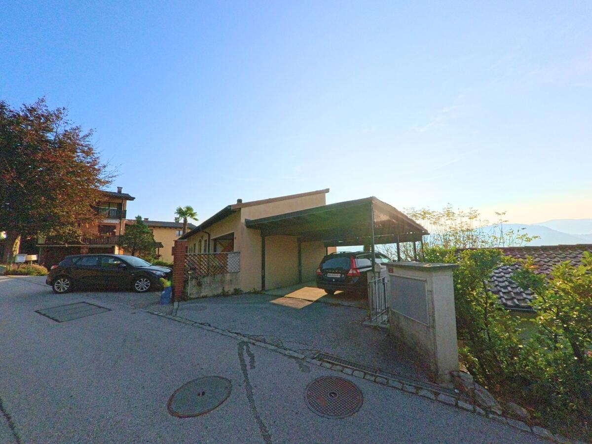 Ein Wohnhaus mit überdachtem Carport liegt an einer ruhigen Quartierstrasse oberhalb des Tals in Vernate (6992, Kanton Tessin) im weichen Abendlicht.