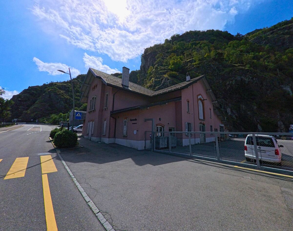 Das Bahnhofgebaeude von Vernayaz (1904, Kanton Wallis) steht mit rosafarbener Fassade am Strassenrand vor steilen bewaldeten Felsen.