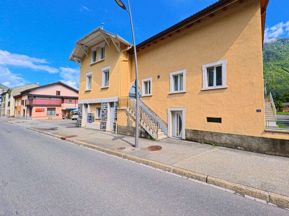 Ein gelbes Wohnhaus mit Steintreppe steht in Vernayaz (1904, Kanton Wallis) an einer Strasse mit weiteren farbigen Gebaeuden.