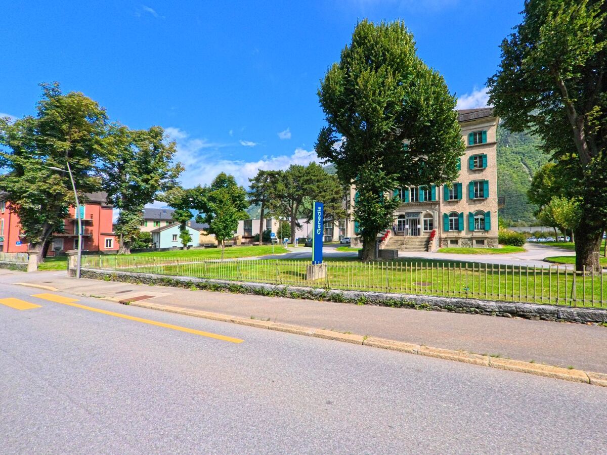 Das Gemeindehaus von Vernayaz (1904, Kanton Wallis) mit gruener Fensterlaeden steht zwischen grossen Baeumen an einer gepflegten Strasse.