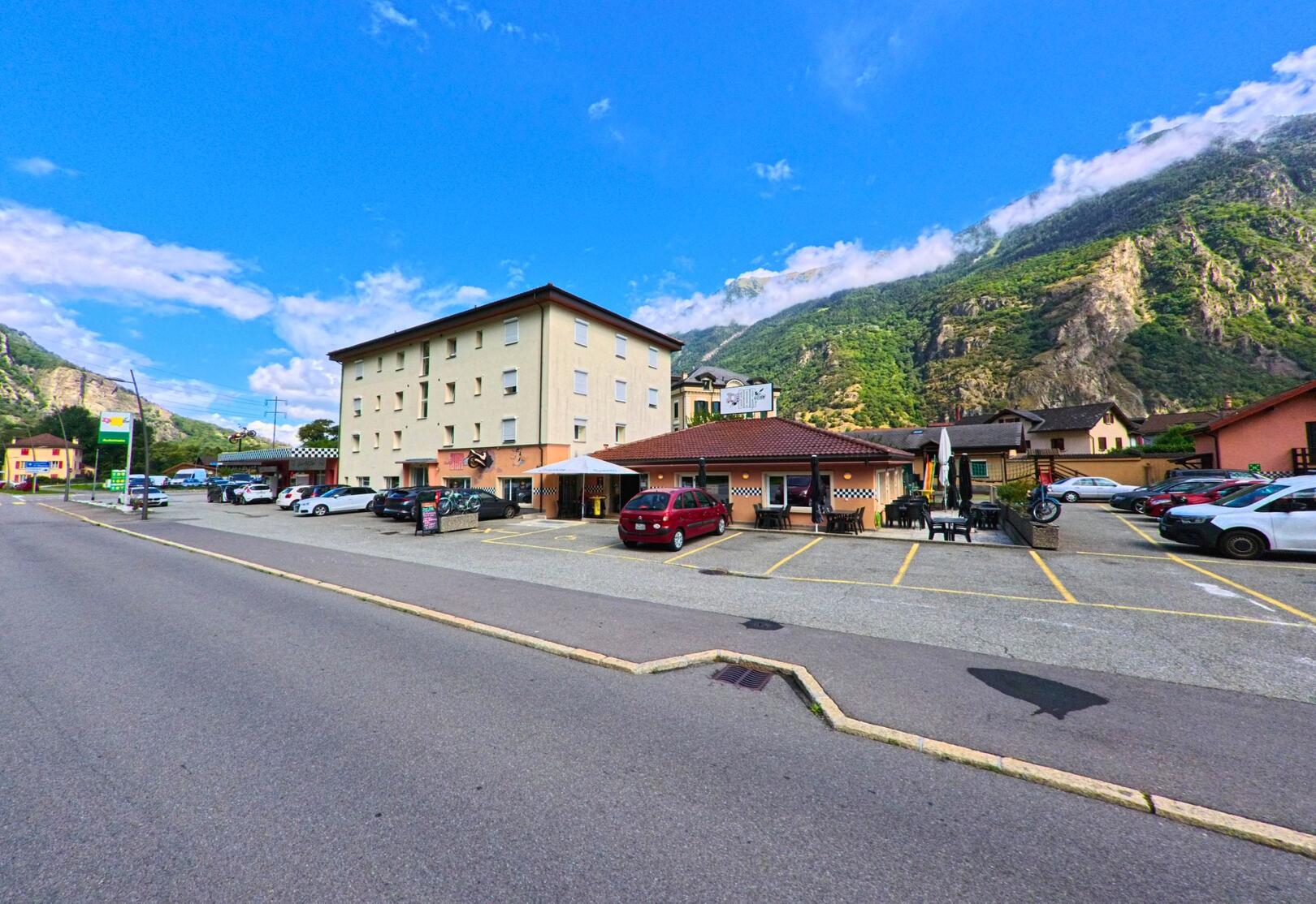 Vor einem Hotel mit Restaurant in Vernayaz (1904, Kanton Wallis) stehen mehrere Autos, dahinter erheben sich steile bewaldete Felswaende.