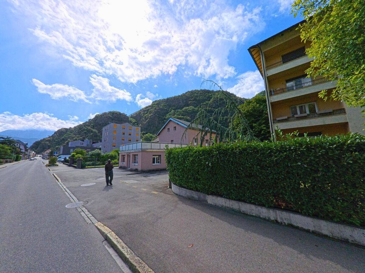 Wohnhaeuser verschiedener Bauweise stehen in Vernayaz (1904, Kanton Wallis) entlang einer Strasse mit Blick auf bewaldete Huegel.