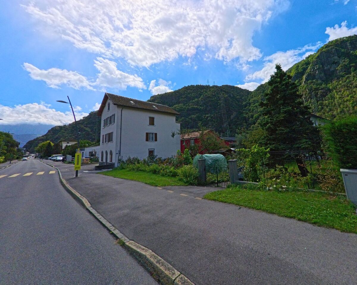 Ein weisses Wohnhaus mit Garten liegt in Vernayaz (1904, Kanton Wallis) an der Strasse, im Hintergrund erheben sich dicht bewaldete Huegel.