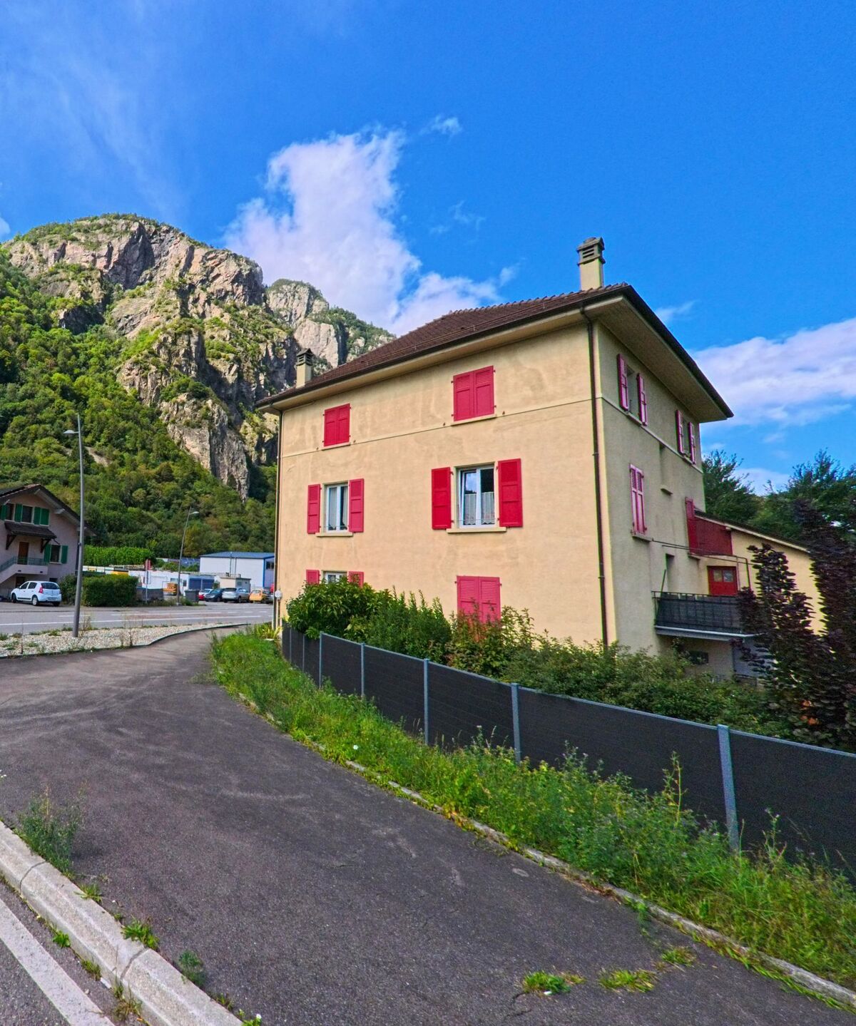 Ein Wohnhaus mit roten Fensterlaeden steht in Vernayaz (1904, Kanton Wallis) vor steilen Felswaenden und gruener Vegetation.