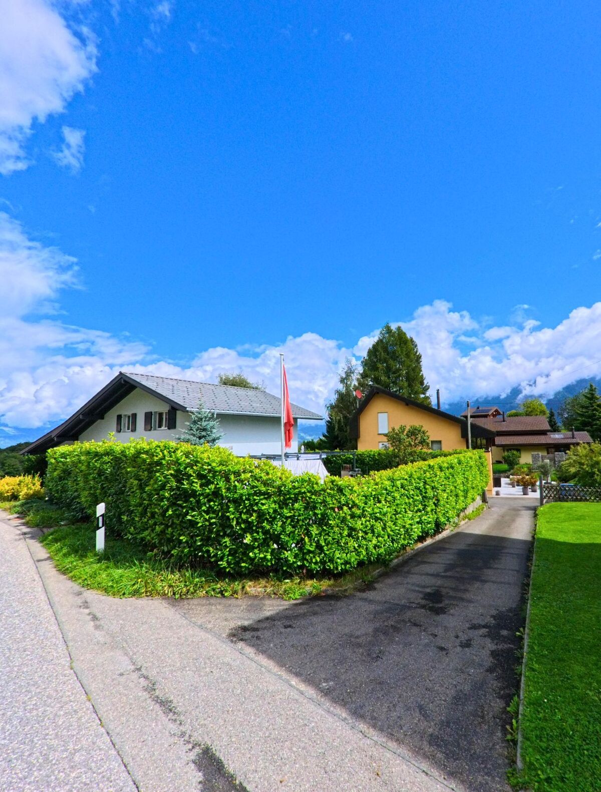 In Vérossaz (1891, Kanton Wallis) fuehrt eine schmale Einfahrt zwischen gruener Hecke zu gepflegten Wohnhaeusern mit Garten.