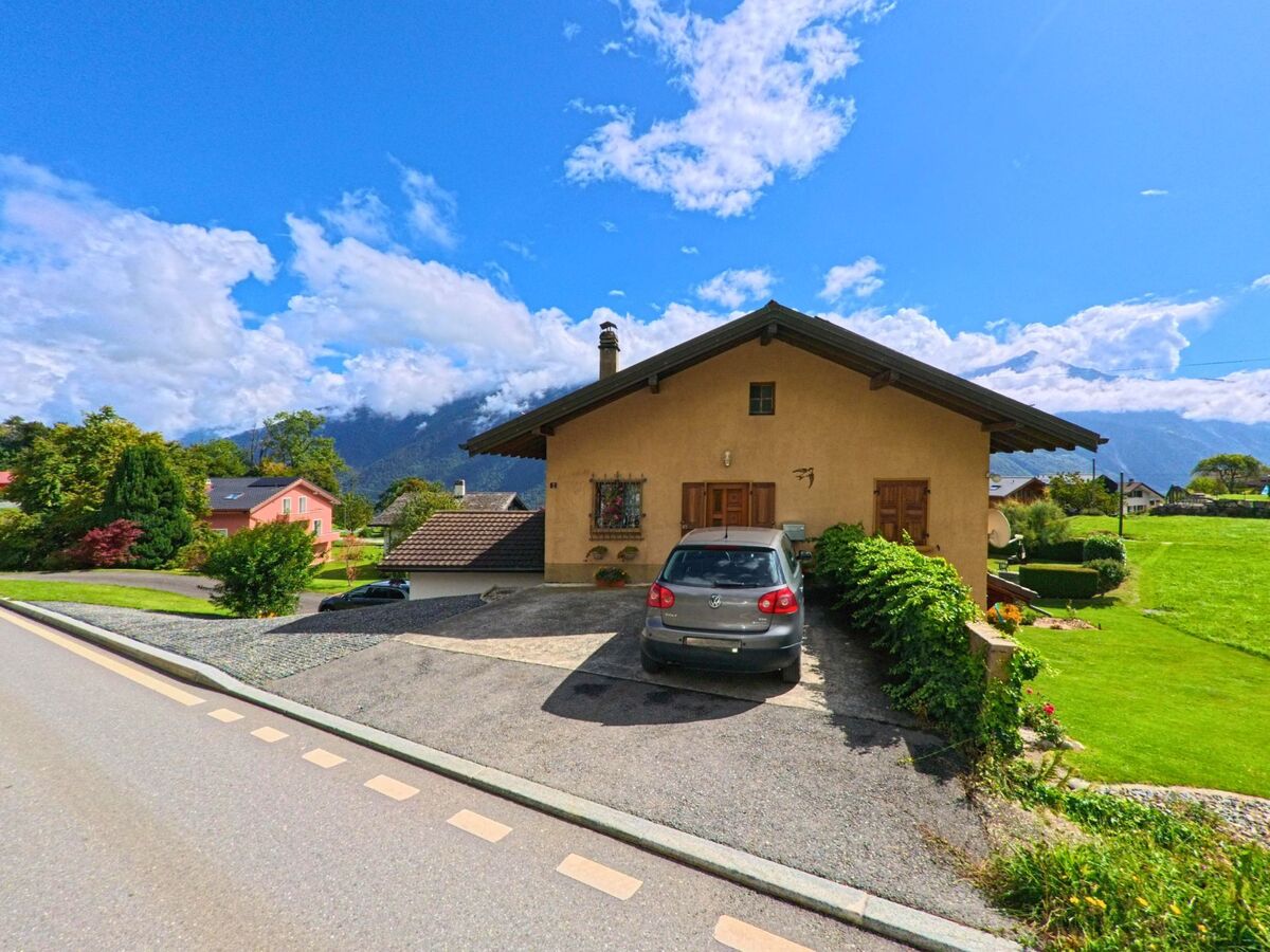 Ein Einfamilienhaus mit geparktem Auto steht in Vérossaz (1891, Kanton Wallis) umgeben von Gruenflaechen und Bergen im Hintergrund.