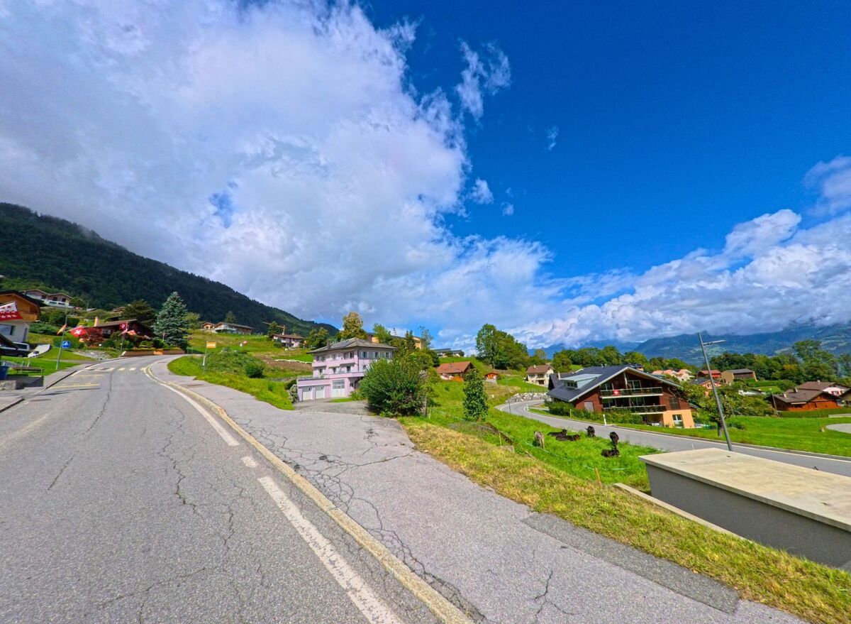 Oberhalb von Vérossaz (1891, Kanton Wallis) liegen Wohnhaeuser am Hang, waehrend eine kurvige Strasse durch die gruenen Wiesen fuehrt.