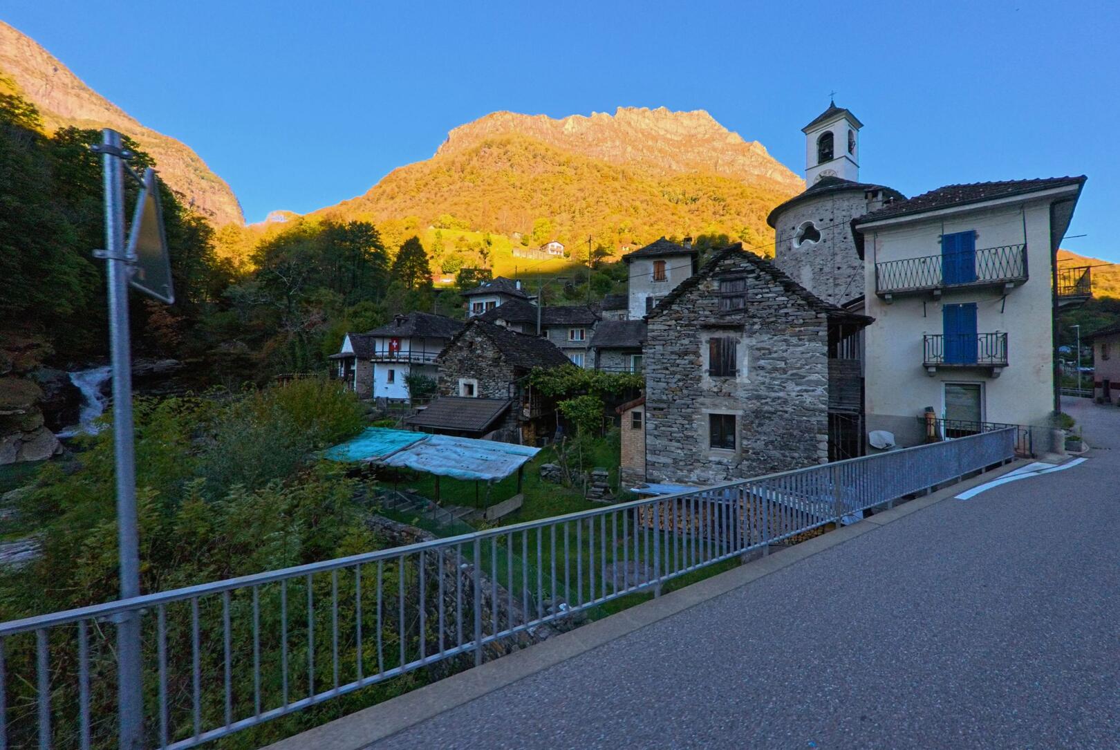 Im Abendlicht liegt das Dorf Verzasca (6637, Kanton Tessin) mit alten Steinhäusern und einer kleinen Kirche am Hang, während im Hintergrund ein beleuchteter Berg aufragt.