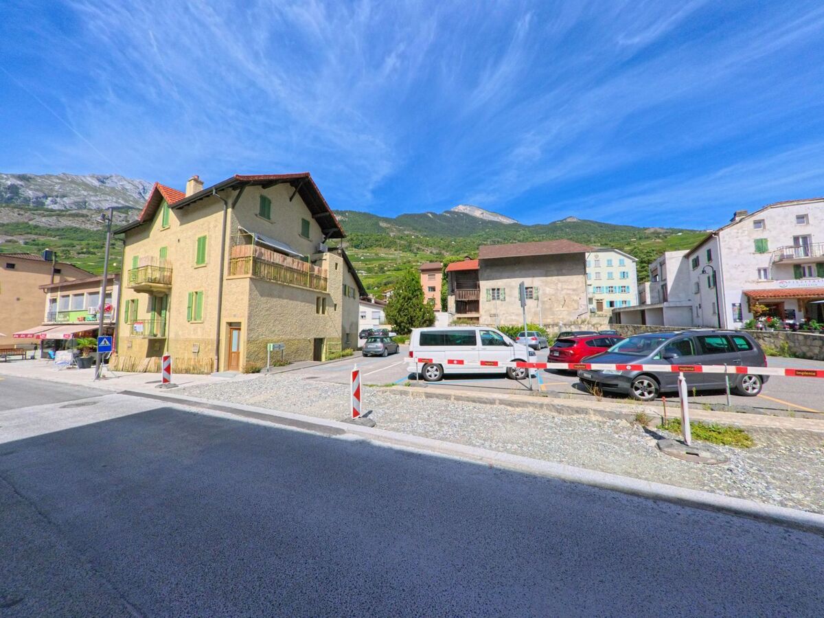 Im Dorfzentrum von Vetroz (1963, Kanton Wallis) stehen traditionelle Wohnhaeuser neben einem Parkplatz mit Fahrzeugen.
