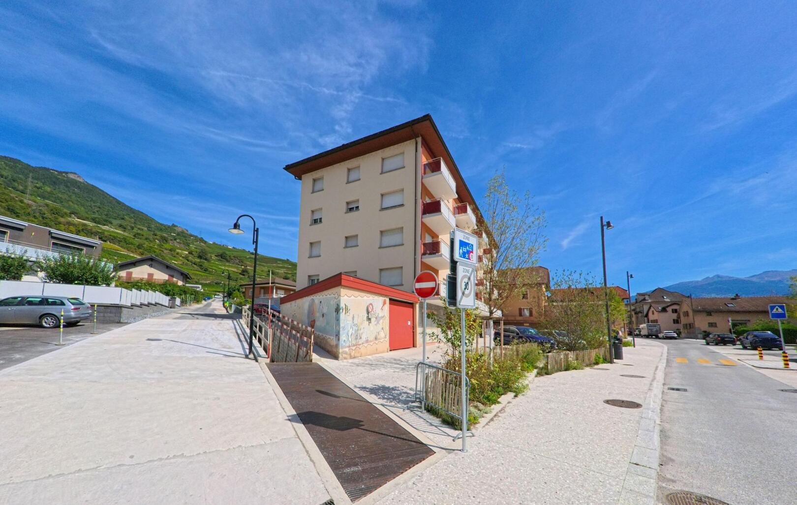 Ein mehrstoekiges Wohnhaus praegt in Vetroz (1963, Kanton Wallis) eine Strassenszene mit Gehwegen und angrenzenden Parkplaetzen.