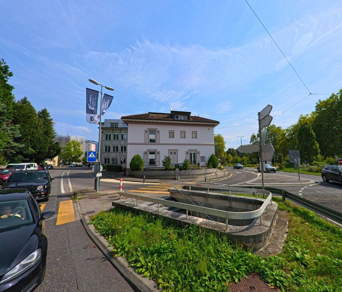Ein weisses Wohnhaus in Vevey (1800, Kanton Waadt) steht an einer mehrspurigen Kreuzung mit Fussgaengeruebergaengen und Gruenstreifen.